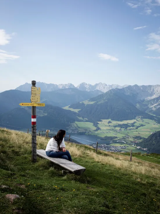 Frau auf Bank neben Wanderwegweiser mit Panoramablick auf die Alpen und das Tal