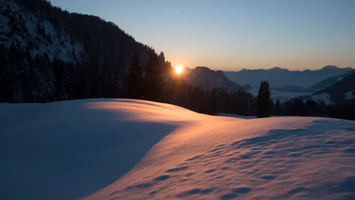 Sunset over snowy alpine landscape with forested hills and distant mountain range