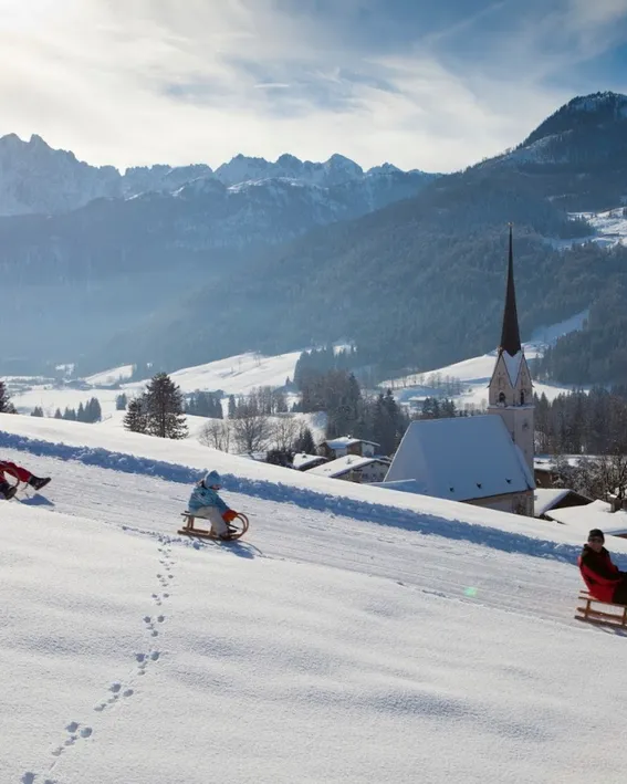 People sledding on a snowy hillside with alpine village and mountains in the background