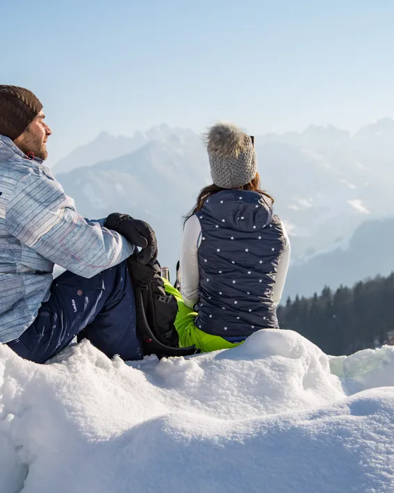 Paar in Winterkleidung sitzt auf verschneitem Hügel mit Blick auf Alpenlandschaft