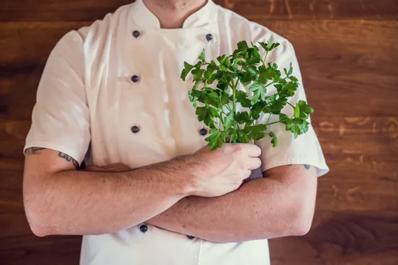 Chef in white uniform holding fresh parsley against wooden background