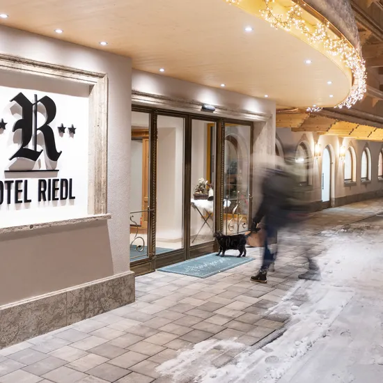 Eingang des Hotel Riedl mit Winterbeleuchtung, schneebedecktem Boden und ankommendem Gast mit Hund