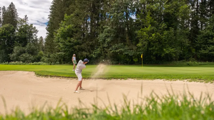 Golfer hitting ball from sand bunker toward green on a forest-lined course