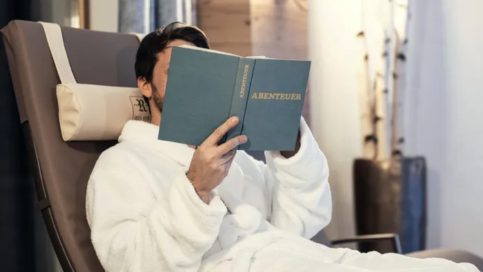 Mann im Bademantel entspannt auf Liege und liest ein Buch mit dem Titel Abenteuer