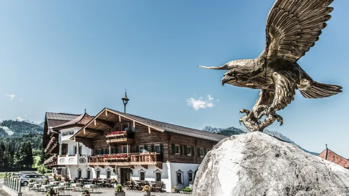 Bronzene Adlerstatue auf Felsen vor Alpenhotel mit Außenterrasse und Bergblick