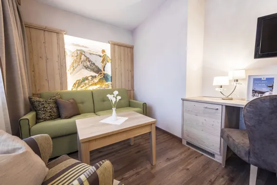 Gemütliches Hotelzimmer mit grünem Sofa, Holzmöbeln und Bergkunst an der Wand