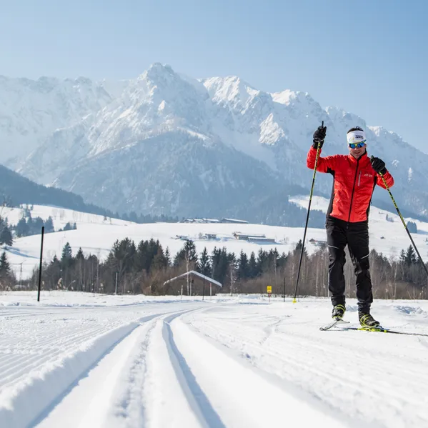 Langläufer in roter Jacke auf präparierter Loipe vor verschneiten Alpenbergen