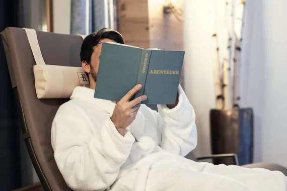 Mann im Bademantel entspannt auf Liege und liest ein Buch mit dem Titel Abenteuer