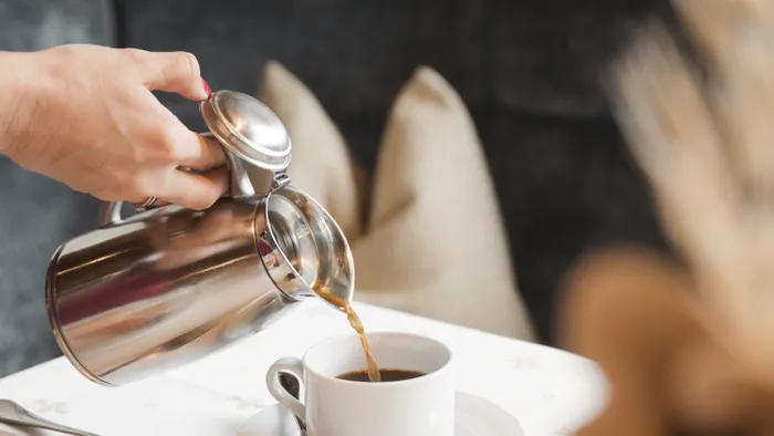 Person gießt Kaffee aus silberner Kanne in eine weiße Tasse auf einem Esstisch