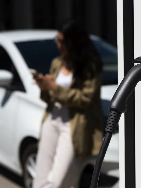 Elektroauto an Ladestation, während Frau Smartphone benutzt
