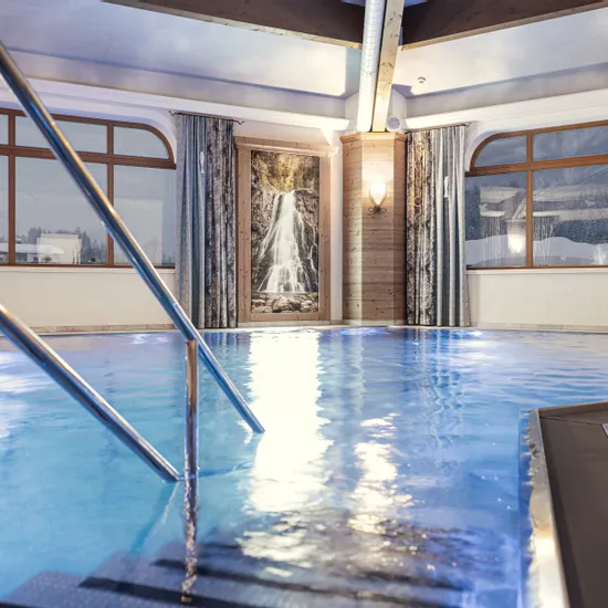 Hotelhallenbad mit warmer Beleuchtung, Wasserfallbild und Bergblick durch große Fenster