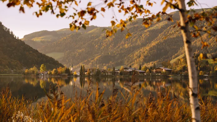 Herbstlicher See mit Alpenort, Kirche und bewaldeten Bergen im warmen Sonnenuntergangslicht