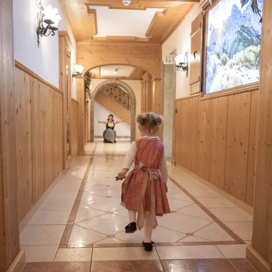 Kleines Mädchen im Dirndl geht den Flur eines Alpenhotels zur Frau hinunter