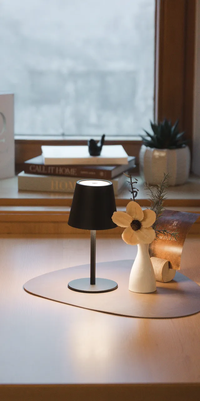 Gemütlicher Raum mit Holztisch, Lampe, Vase mit Blume, Büchern und Deko am Fenster