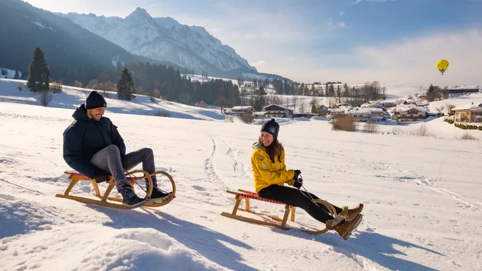 Lächelndes Paar beim Rodeln auf verschneitem Feld vor Alpenkulisse und Bergdorf