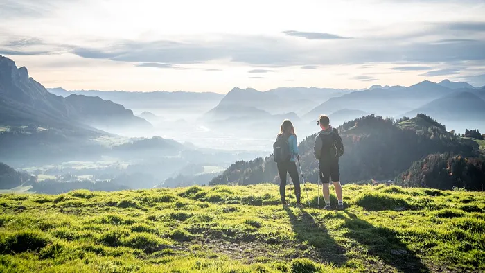 Hikers with backpacks standing on grassy hill overlooking scenic mountain valley at sunrise