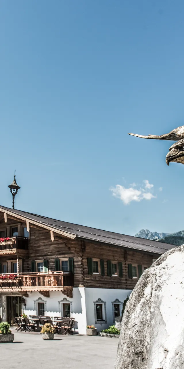Bronzene Adlerstatue auf Felsen vor Alpenhotel mit Außenterrasse und Bergblick