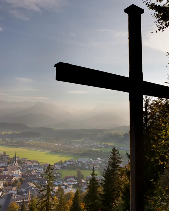 Holzkreuz auf Hügel mit Blick auf alpines Dorf und Berge bei Sonnenaufgang