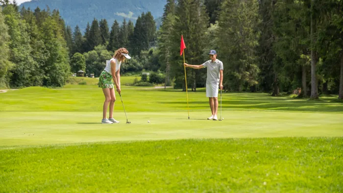 Woman putting on green while man holds flagstick on scenic golf course