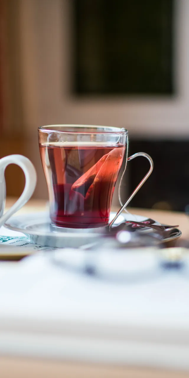 Glastasse mit schwarzem Tee und Teekanne auf Tisch neben Buch und Lesebrille