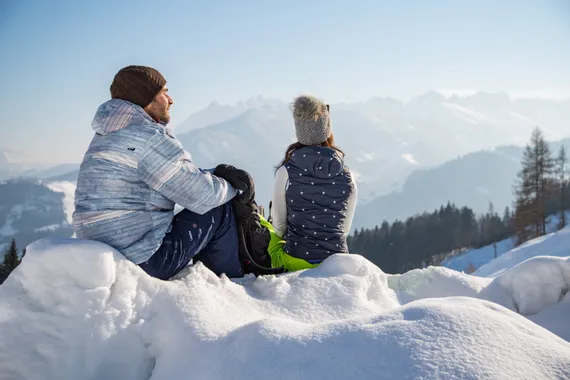 Paar in Winterkleidung sitzt auf verschneitem Hügel mit Blick auf Alpenlandschaft