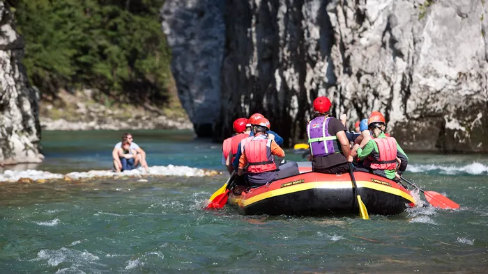 Group rafting on a mountain river with rocky cliffs and a couple sitting on a riverbank
