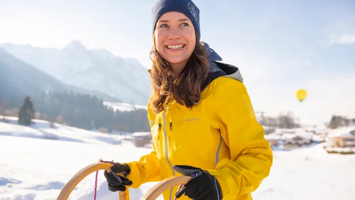 Smiling woman in yellow jacket holding sled in snowy alpine landscape