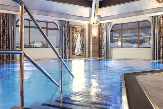 Hotelhallenbad mit warmer Beleuchtung, Wasserfallbild und Bergblick durch große Fenster