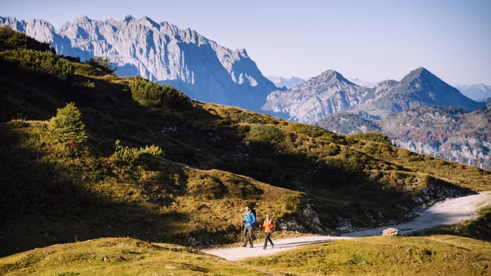 Two hikers walking on a mountain trail with alpine peaks in the background