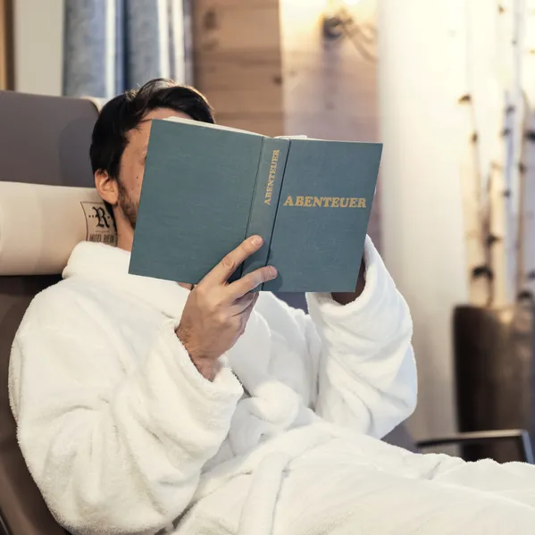 Mann im Bademantel entspannt auf Liege und liest ein Buch mit dem Titel Abenteuer