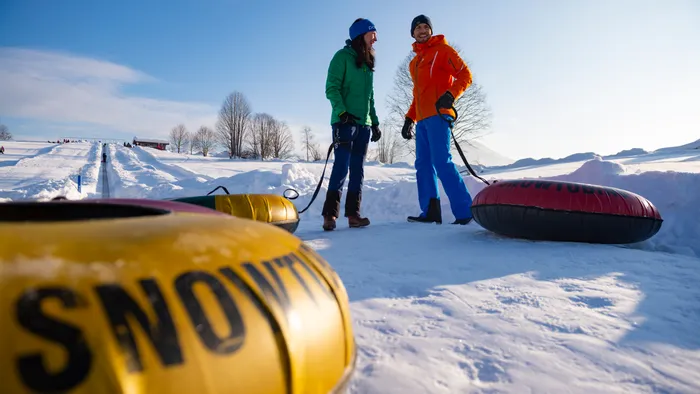 Two people with snow tubes standing on a snowy hill under clear blue sky