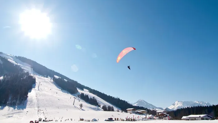 Paraglider soaring above snowy ski resort with mountains and clear blue sky