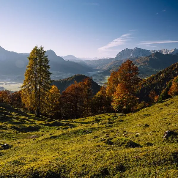 Autumn mountain landscape with green meadow, colorful trees, and alpine peaks under a clear blue sky