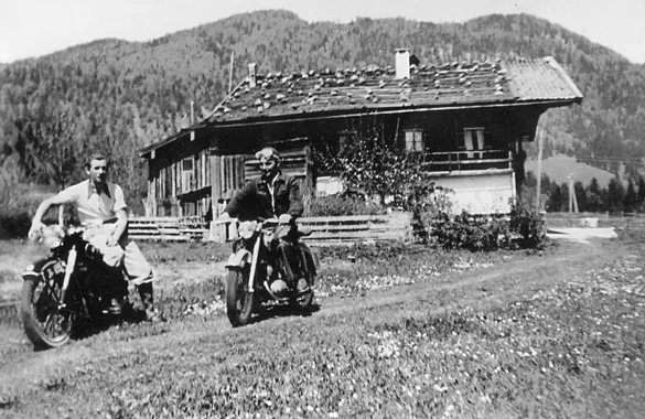 Zwei Männer auf Oldtimer-Motorrädern vor einer Berghütte in alpiner Landschaft