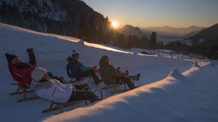 People sledding on a snowy mountain trail at sunset with alpine landscape in the background