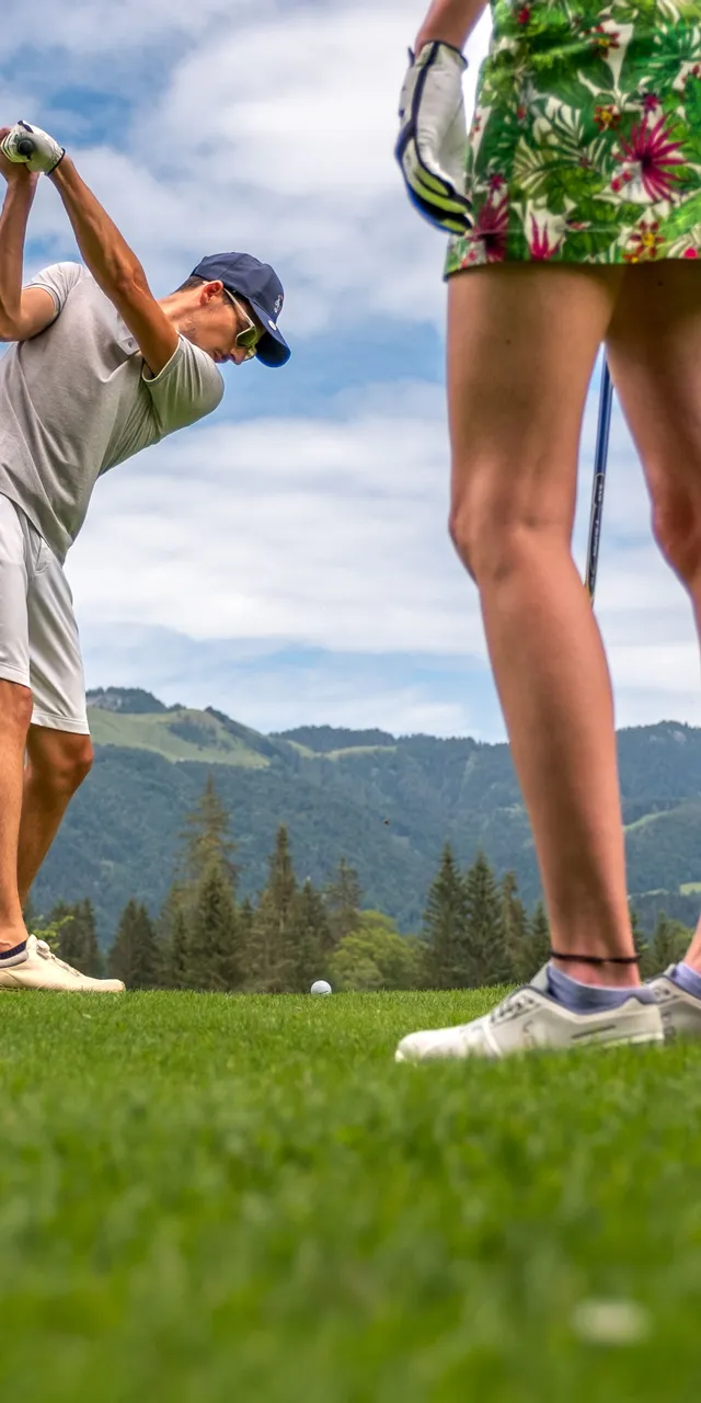Man swinging golf club on green fairway with mountains and forest in background