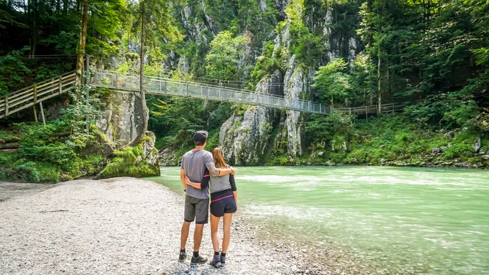 Paar umarmt sich an einem türkisfarbenen Fluss, Wald und Seilbrücke im Tal