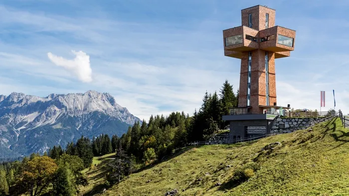 Modern wooden viewing tower on grassy hill with alpine mountains in the background
