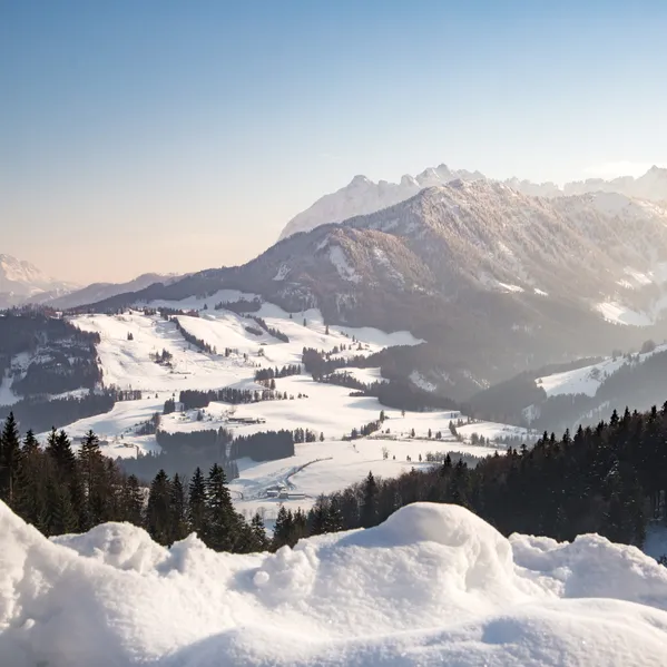 Snow-covered alpine landscape with forested hills and distant mountain range at sunrise