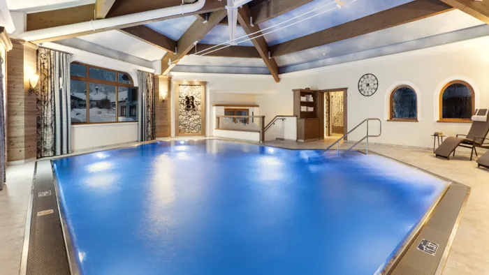 Indoor hotel pool with glowing blue water, wooden beams, lounge chairs and winter view outside
