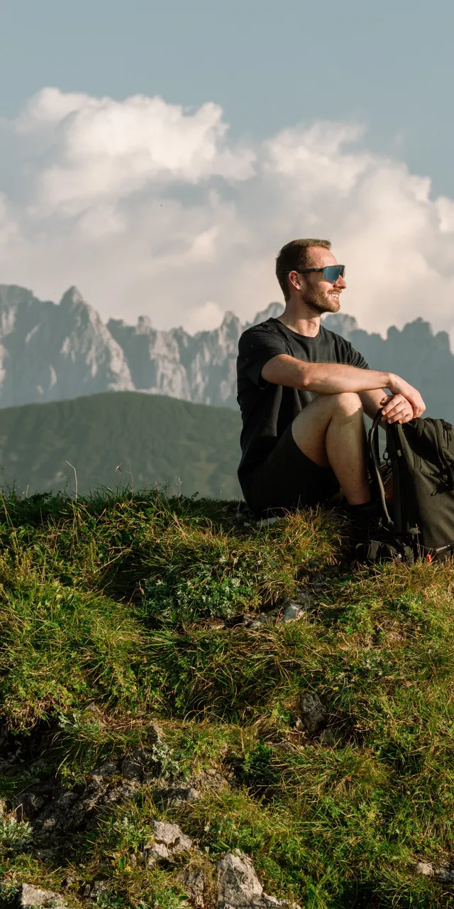 Man sitting on grassy mountain peak with backpack, alpine landscape in the background