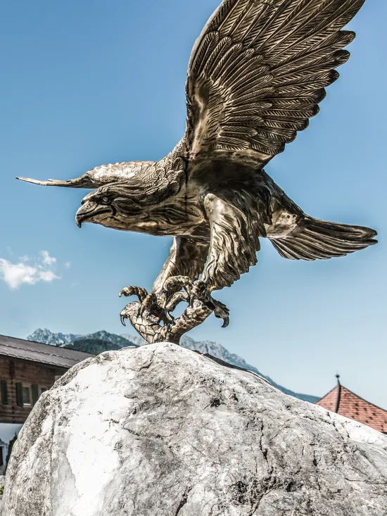 Bronzene Adlerstatue auf Felsen vor Alpenhotel mit Außenterrasse und Bergblick