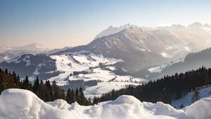 Snow-covered alpine landscape with forested hills and distant mountain range at sunrise
