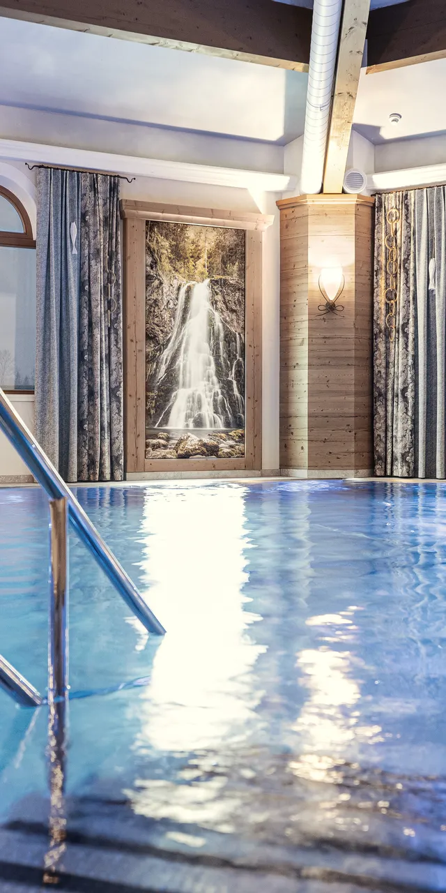 Hotelhallenbad mit warmer Beleuchtung, Wasserfallbild und Bergblick durch große Fenster
